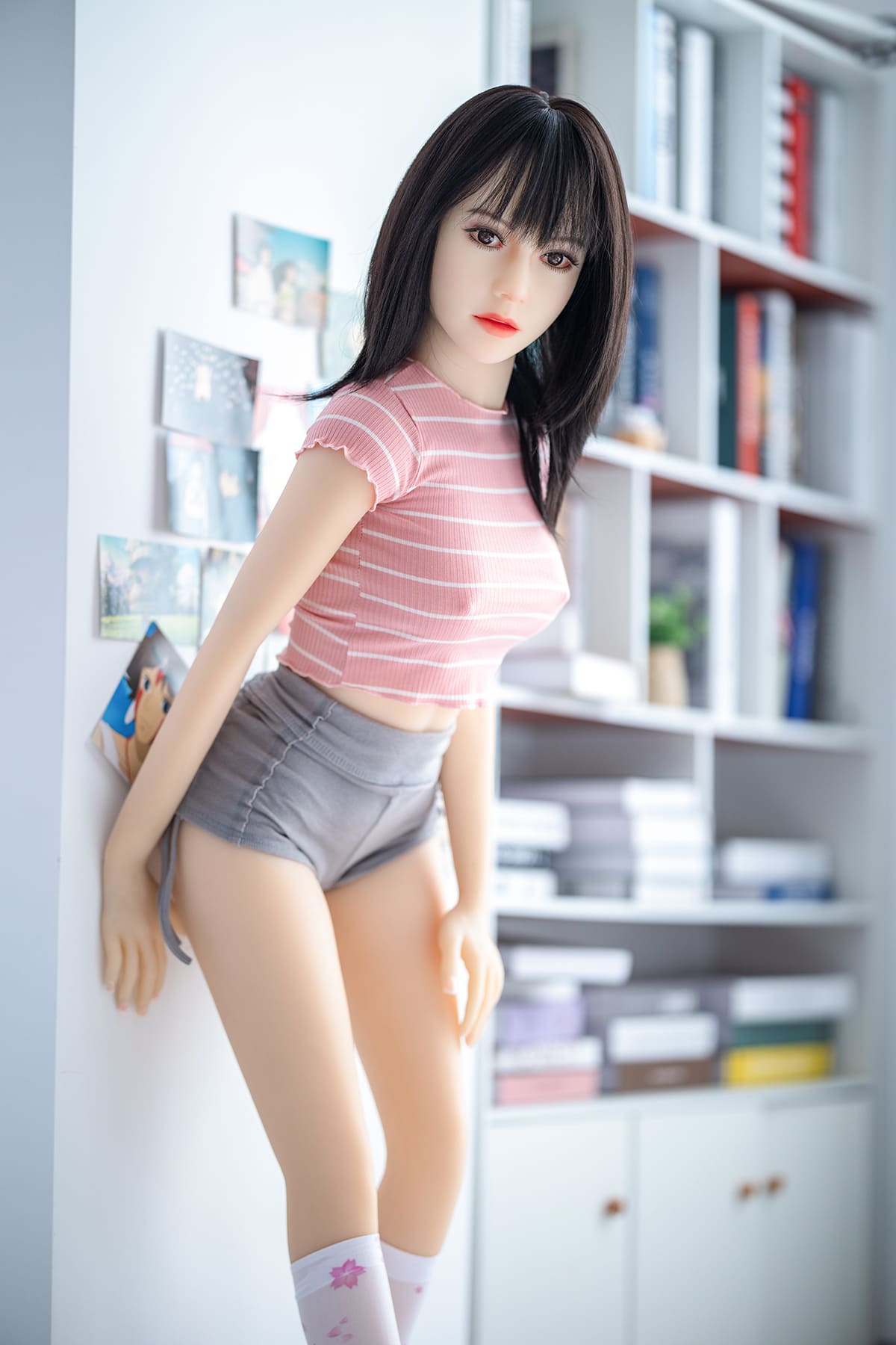 Young female sex doll wearing pink short sleeve and grey shorts in library